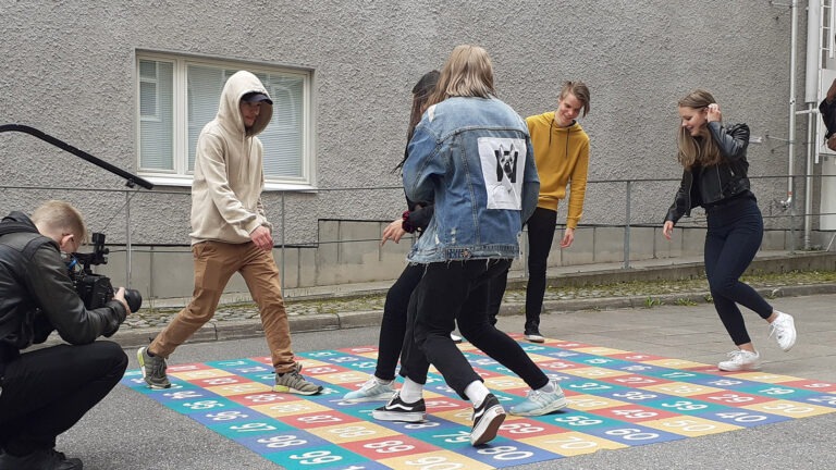 ryhmä nuoria pelaa asfalttiin maalattua peliä ja kameramies kuvaa heitä