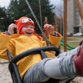 Kuva, jossa nuori lapsi nauraa keinussa. Laidassa näkyy kuinka aikuisen kädet antavat hänelle vauhtia. Lapsella on keltainen takki ja punainen pipo ja taustana on leikkipuisto.
