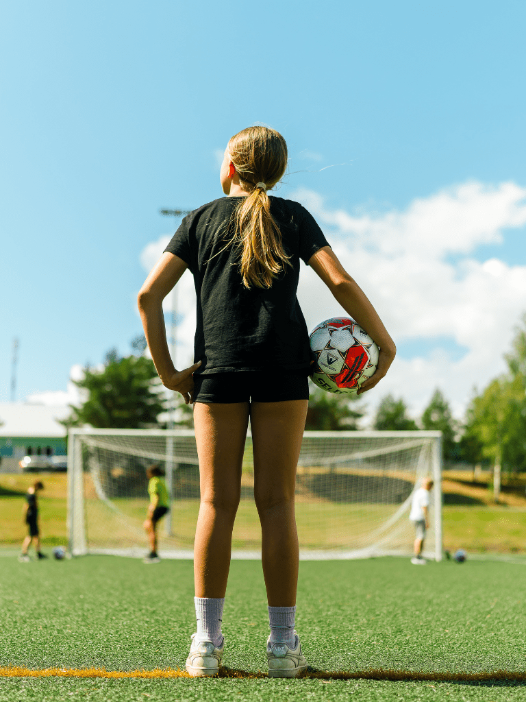 Kuvassa tyttö selin kameraan päin, kainalossa tytöllä on jalkapallo ja hänen edessään on jalkapallomaali.