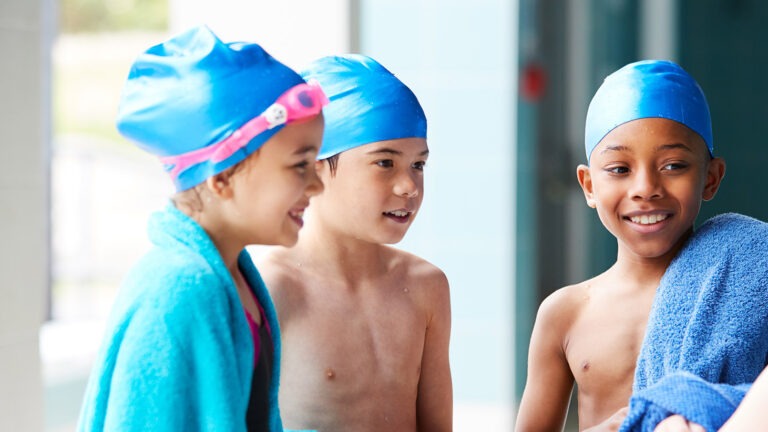 Kuva, jossa näkyy neljä iloista lasta uimapuvuissa. Kaikilla on sinisen uimalakit päässä ja kahdella on pyyhkeet.