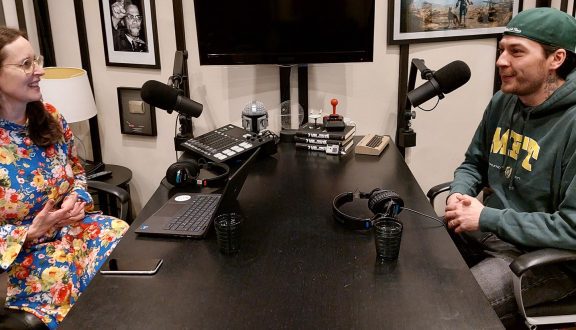 Kuva, jossa näkyy äänitysstudio. Kaksi ihmistä istuu pöydän ääressä mikkien edessä, yksi ruskeahiuksinen nainen silmälaseissa ja kukallisessa mekossa ja yksi mies lippalakki päässä ja vihreä huppari päällä.