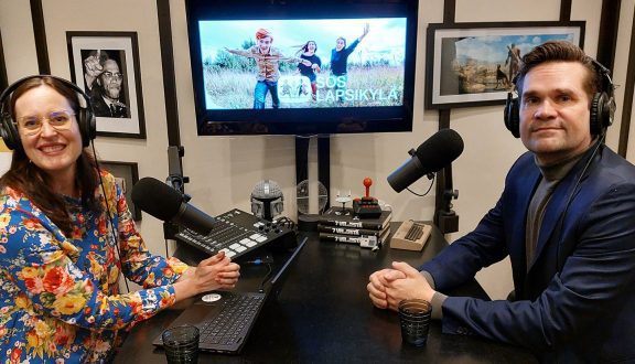 Kuva, jossa kukalliseen mekkoon pukeutunut nainen ja tummansiniseen villapaitaan pukeutunut mies istuvat äänitysstudiossa kuulokkeet korvailla ja katsovat kameraan.