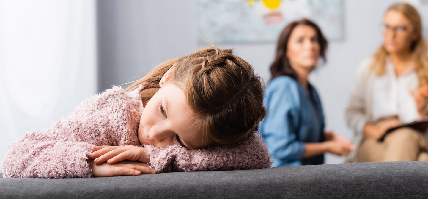 tyttö vaaleanpunaisessa paidassa nojaa päätänsä käsiin, taustalla kaksi aikuista keskustelee