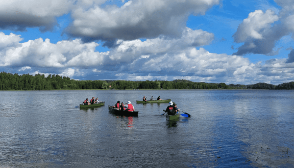 Kuvassa neljä kanoottia kesäisellä järvellä, jokaisessa kanootissa on kolme ihmistä.