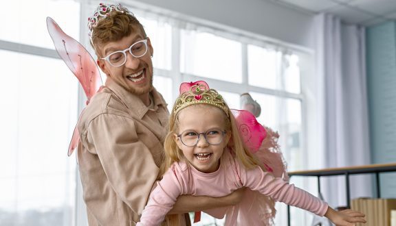 Kuvassa mies kannattelee pientä tyttöä. Molemmat ovat pukeutuneet keijun siipiin ja kruunuihin. Molemmat nauravat kuvassa.