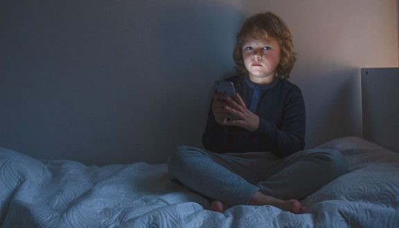 Poika istuu hämärässä huoneessa sängyllään kädessään känykkä