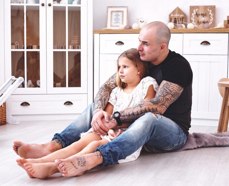 Vaaleasävyinen lapsen huone, jossa on puisia huonekaluja. Pieni vaaleahiuksinen tyttö valkoisessa mekossa. Isä, jolla on tatuointeja halaa häntä. Molemmat katsovat kaukaisuuteen.