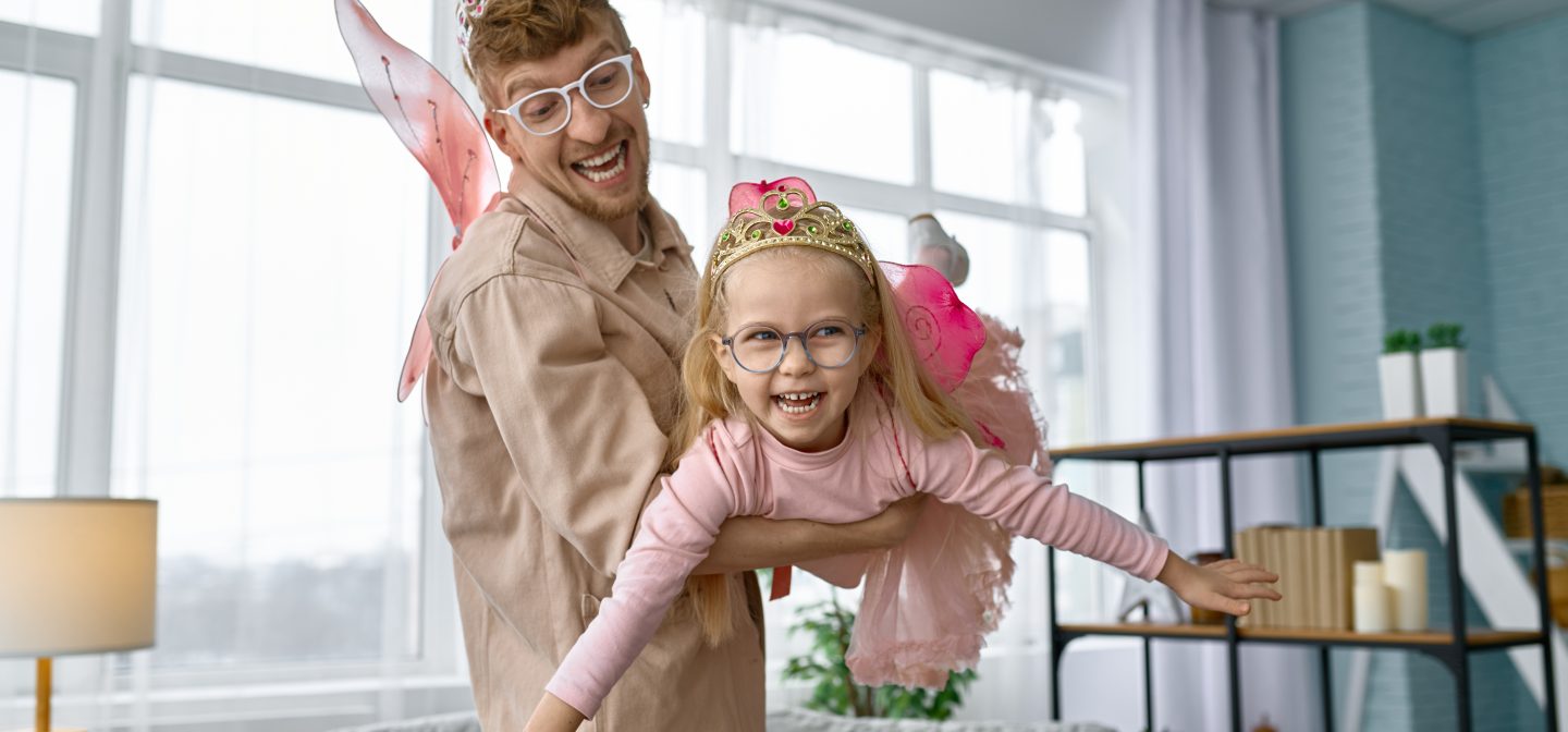 Iloinen isä ja innostunut pieni tytär ovat pukeutuneet keijuiksi. Isä pitää tytärtä sylissä.