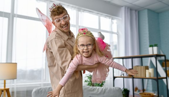Iloinen isä ja innostunut pieni tytär ovat pukeutuneet keijuiksi. Isä pitää tytärtä sylissä.