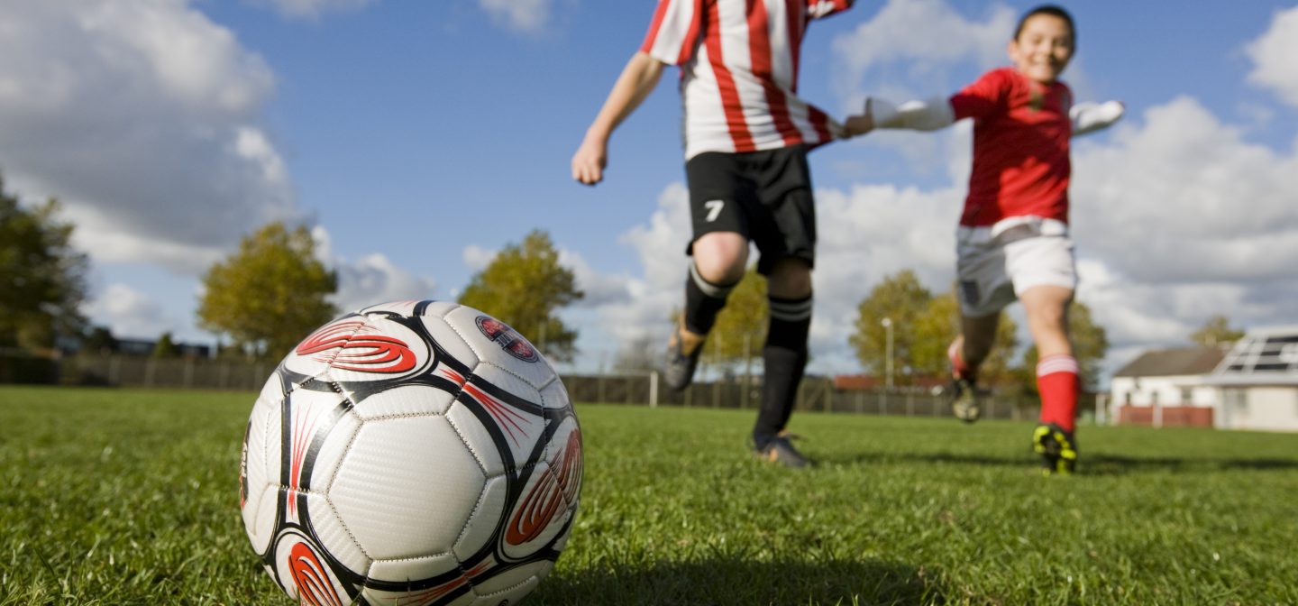 Kaksi lasta juoksee pallon perässä aurinkoisena päivänä nurmikolla.