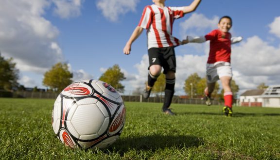 Kaksi lasta juoksee pallon perässä aurinkoisena päivänä nurmikolla.