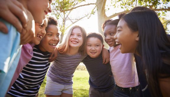Ryhmä kouluikäisiä lapsia seisoo ympyrässä kädet toistensa olkapäillä hymyillen