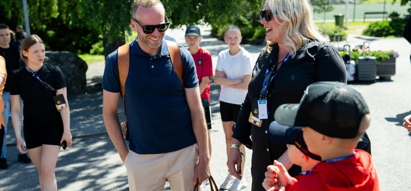 Mika Poutala keskustelee nuoren kanssa hymyillen saapuessaan leirille auringon paisteessa. Kuvassa Mikan vieressä myös nainen ja taka-alalla lapsia jotka näyttävät innostuneelta.