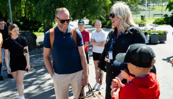 Mika Poutala keskustelee nuoren kanssa hymyillen saapuessaan leirille auringon paisteessa. Kuvassa Mikan vieressä myös nainen ja taka-alalla lapsia jotka näyttävät innostuneelta.