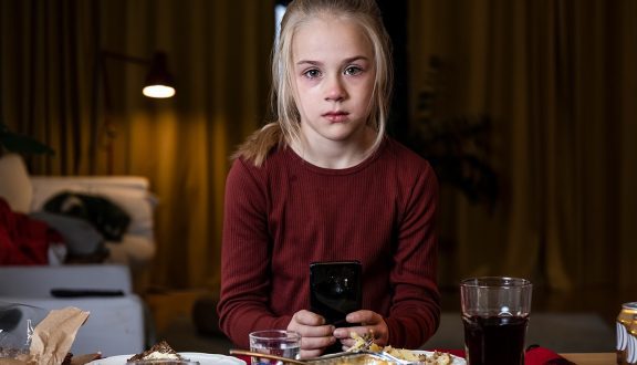 vakava tyttö istuu puhelin kädessä pöydän ääressä ja katsoo kameraan