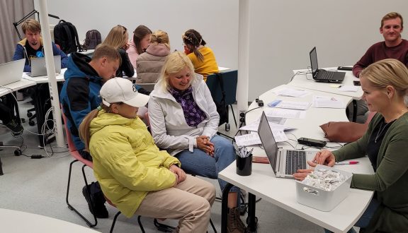 toimistohuone, jossa ihmisä pöytien ympärillä. Sekä aikuisia ja lapsia. Tausalla valkoiset lattiat ja seinät