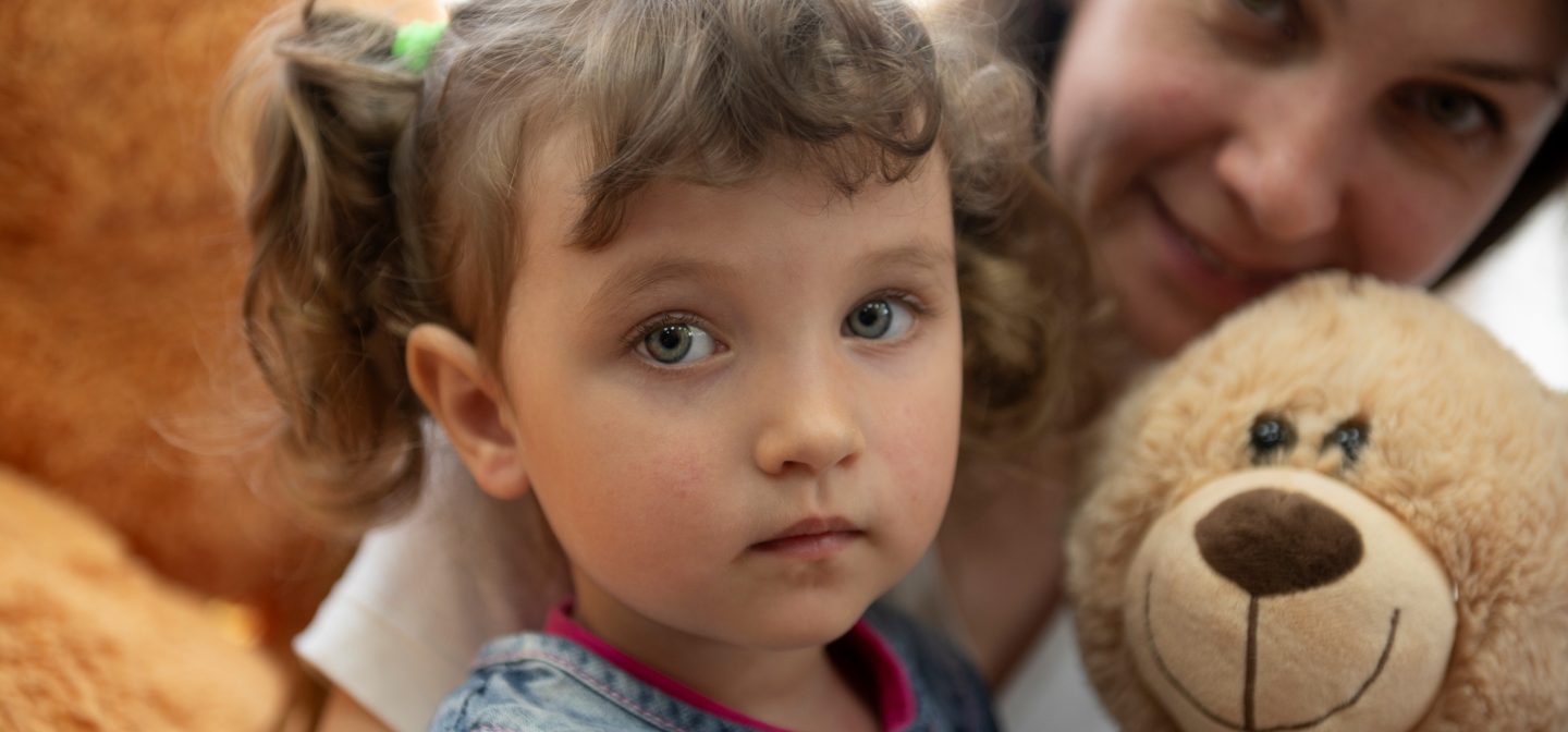 Lapsi katsoo suoraan kameraan nalle kädessä. Äiti on takana.