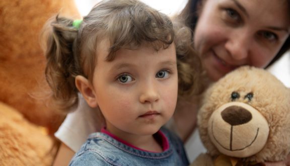 Lapsi katsoo suoraan kameraan nalle kädessä. Äiti on takana.