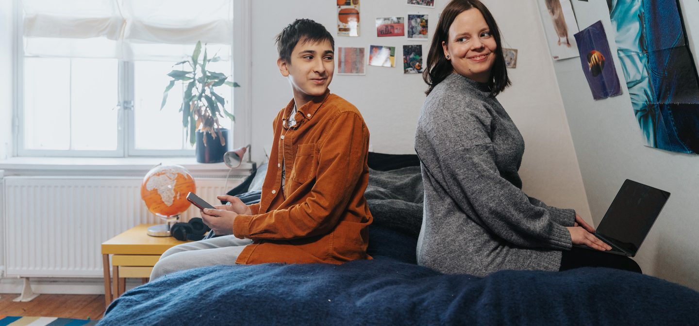 Nuori poika ja nainen istuvat seläkkäin sängyllä, pojalla on kännykkä ja naisella on läppäri.
