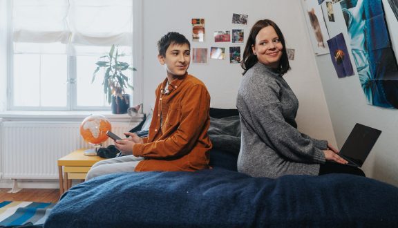Nuori poika ja nainen istuvat seläkkäin sängyllä, pojalla on kännykkä ja naisella on läppäri.