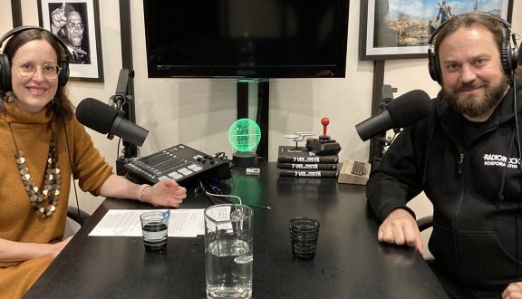 studiossa istuu nainen ruskeassa mekossa ja parrakas mies mikrofonien äärellä