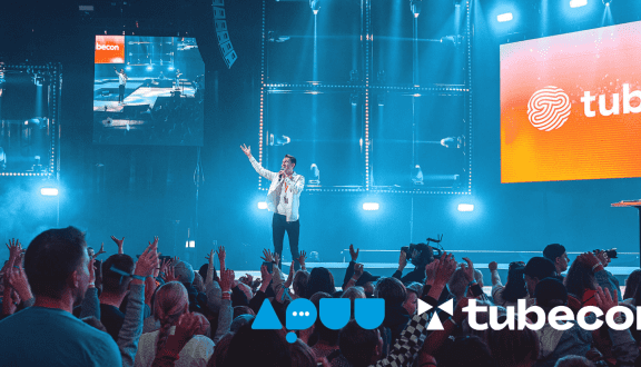 Kuvassa yleisöä Tubeconin lavan edessä, lavalla henkilö puhumassa mikrofoniin. Lavalla screen, jossa teksti "Tubecon".