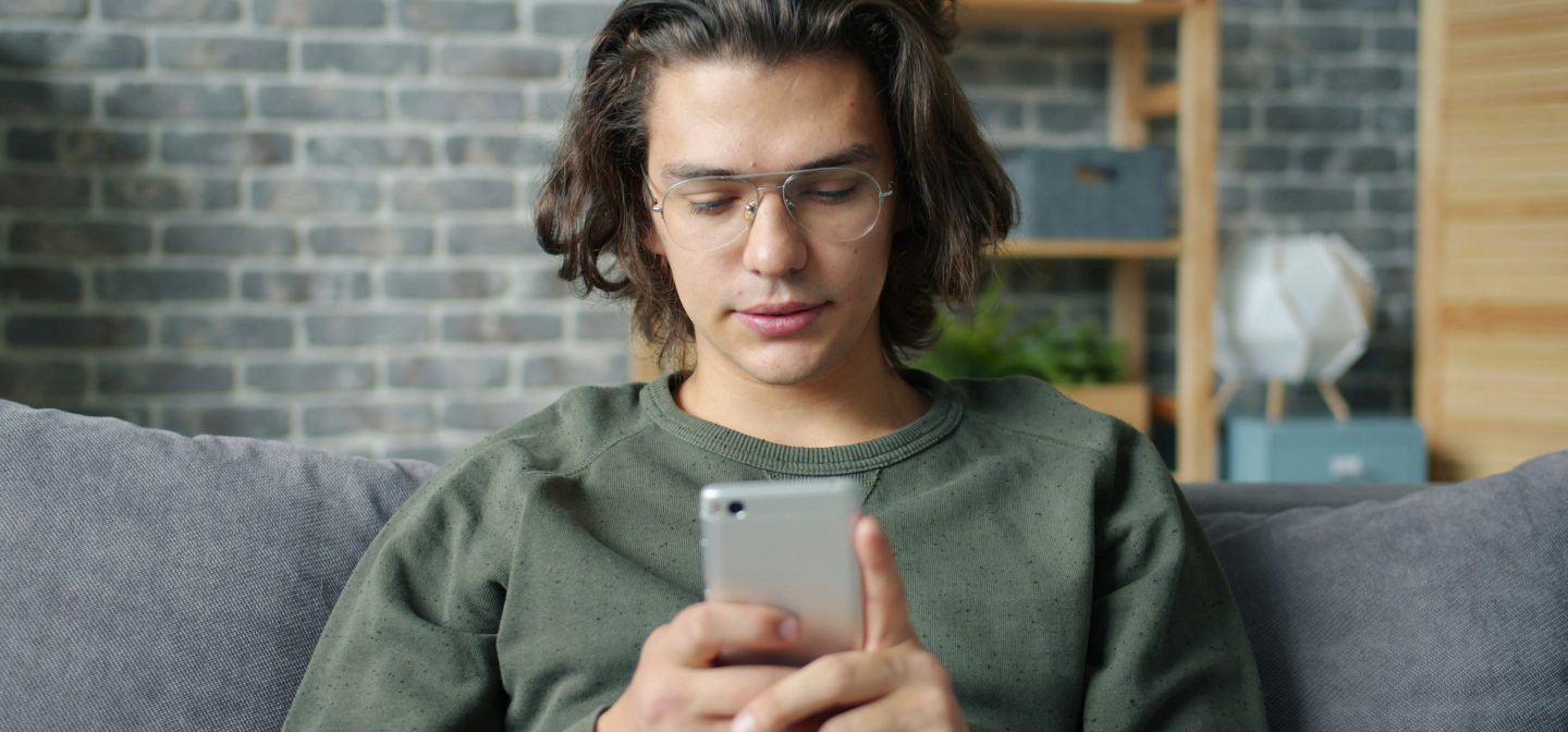 silmälasipäinen poika istuu sohvalla katsoen puhelintaan.