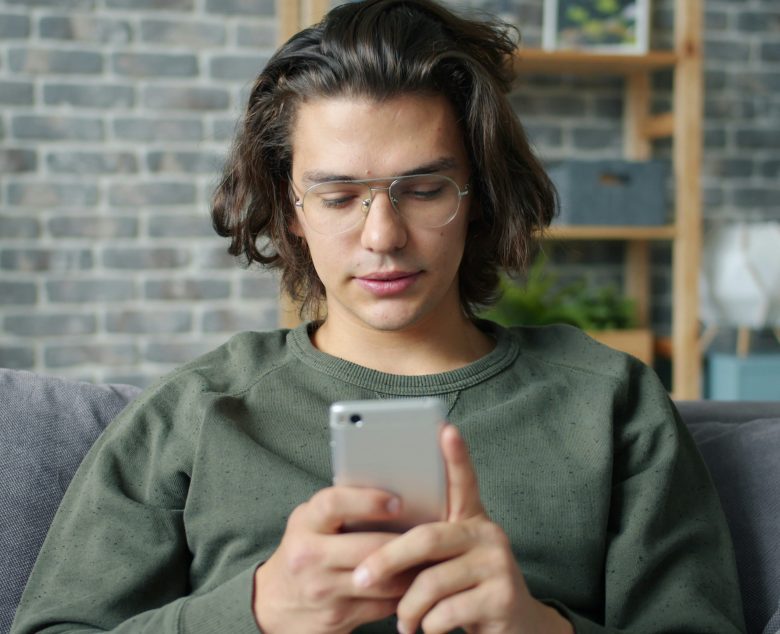 silmälasipäinen poika istuu sohvalla katsoen puhelintaan.